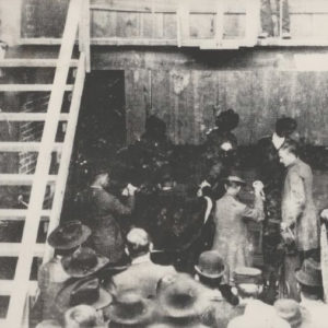 A group of men examining and crowd looking on, with a wooden stairway to a raised platform