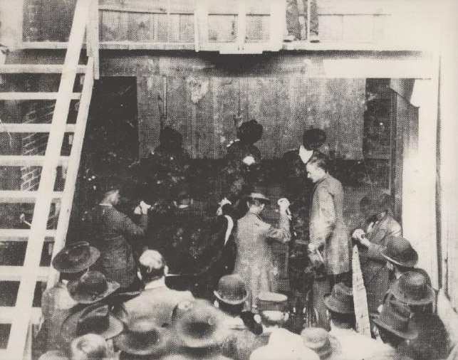 A group of men examining and crowd looking on, with a wooden stairway to a raised platform