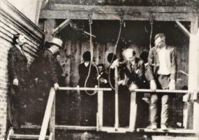 White men in suits inspecting nooses at gallows with hooded men waiting behind them