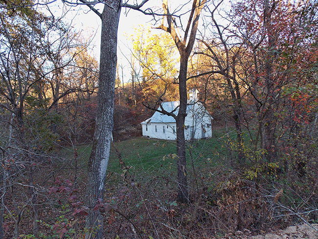 Single-story church building with cupola in grassy valley as seen through trees
