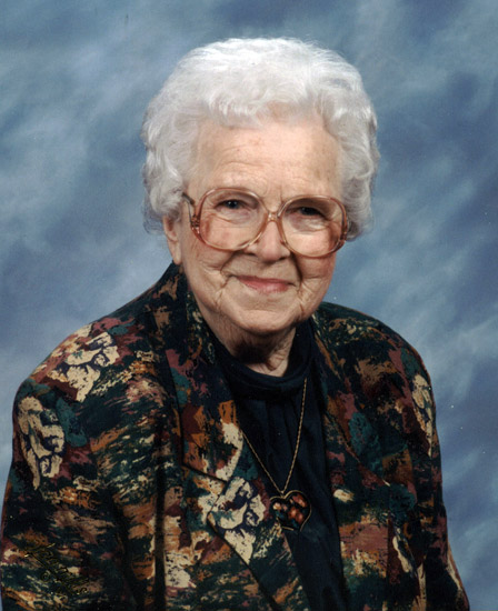 Old white woman with glasses in multicolored jacket and heart shaped pendent necklace with blue background
