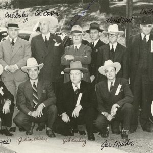 Group of white men in cowboy hats and suits posing together with cars parked behind them