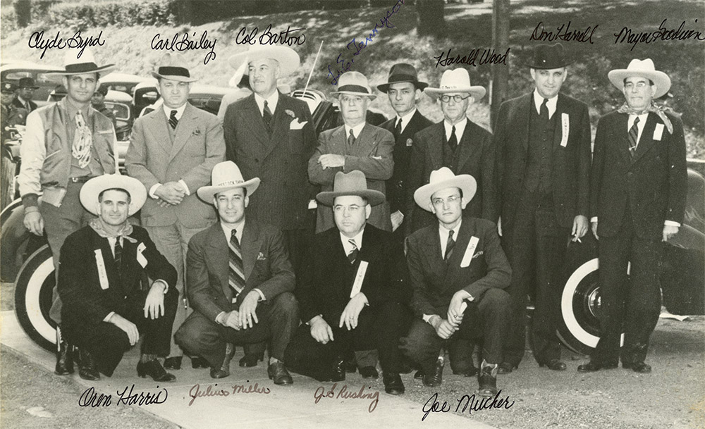 Group of white men in cowboy hats and suits posing together with cars parked behind them