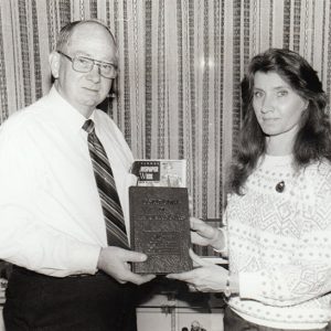 White man in glasses and tie presenting book to white woman