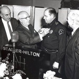 Bald white men in suits giving an award to white man in police uniform at lectern