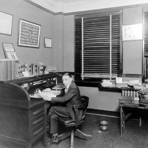 White man sitting at roll-top desk in office with framed diplomas on the wall