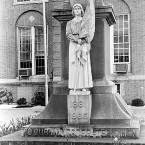 Statue of woman holding flag labeled "to our Confederate women" located in front of tall stone obelisk with brick building in background