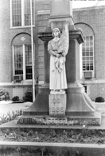 Statue of woman holding flag labeled "to our Confederate women" located in front of tall stone obelisk with brick building in background