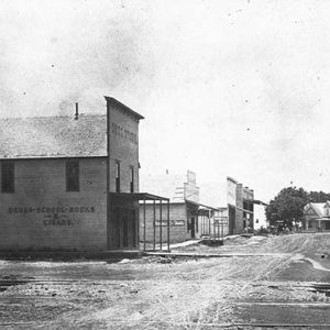 Storefronts with covered porches on dirt road with multistory house in the background and train tracks with hand cart in the foreground