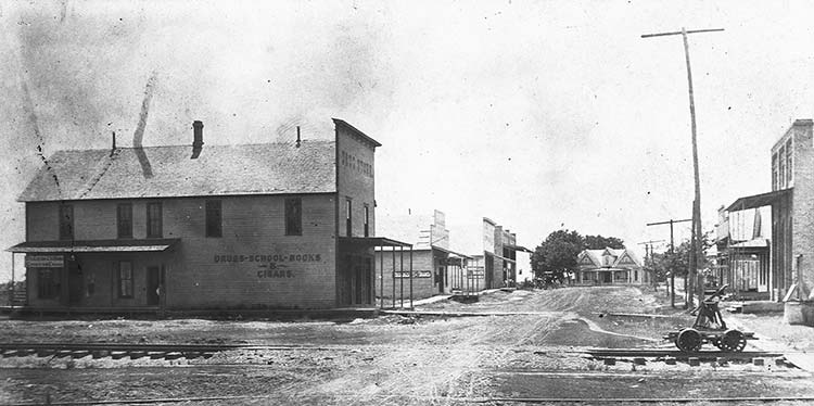 Storefronts with covered porches on dirt road with multistory house in the background and train tracks with hand cart in the foreground