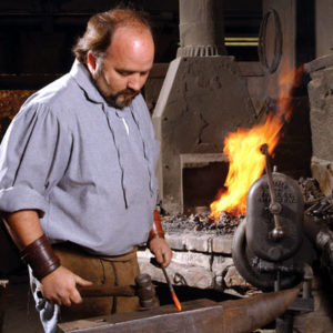 Balding white man preparing to hammer a glowing piece of metal on an anvil