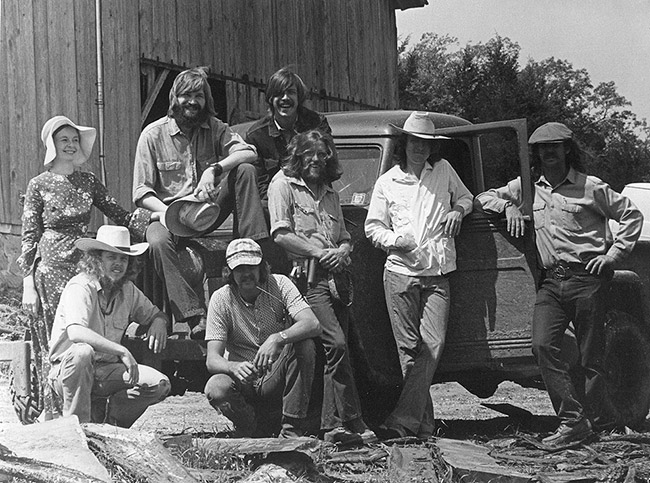Group of long haired men and woman posing with truck and barn in the background