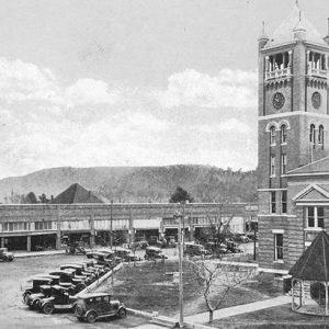 Cars parked outside multistory building with clock tower and gazebo across from line of storefronts on town street
