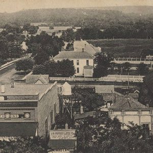 Looking from an elevated vantage point out on brick storefront and agricultural buildings with multistory house in the background