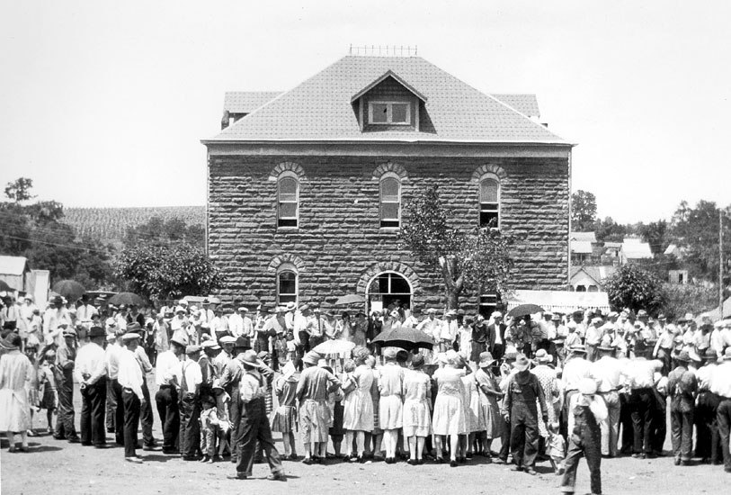 Two-story brick building with arched windows surrounded by a crowd of people, some with umbrellas