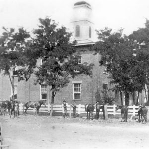 Two-story building with bell tower with trees and people with horses and wagon