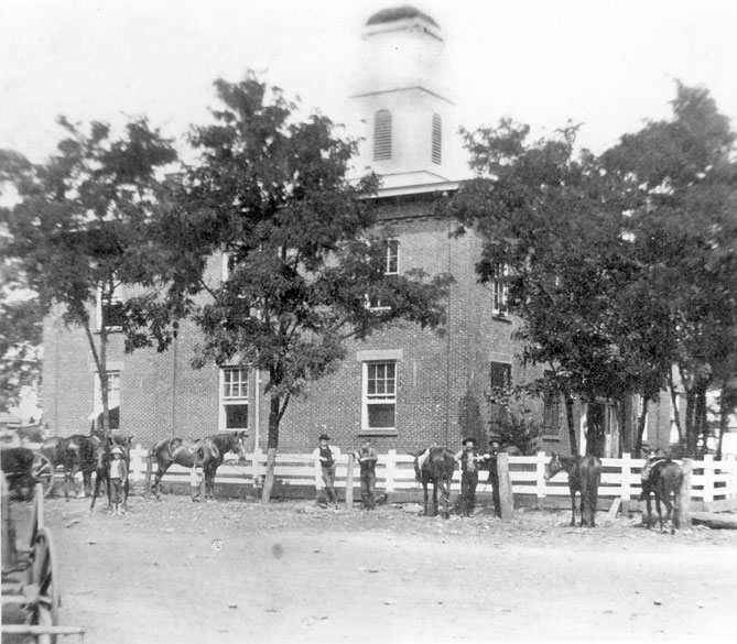 Two-story building with bell tower with trees and people with horses and wagon