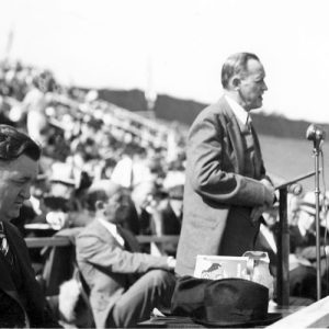 White man speaking at a lectern with crowd behind him