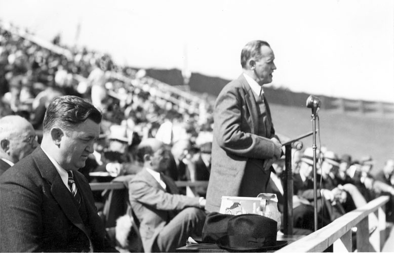 White man speaking at a lectern with crowd behind him