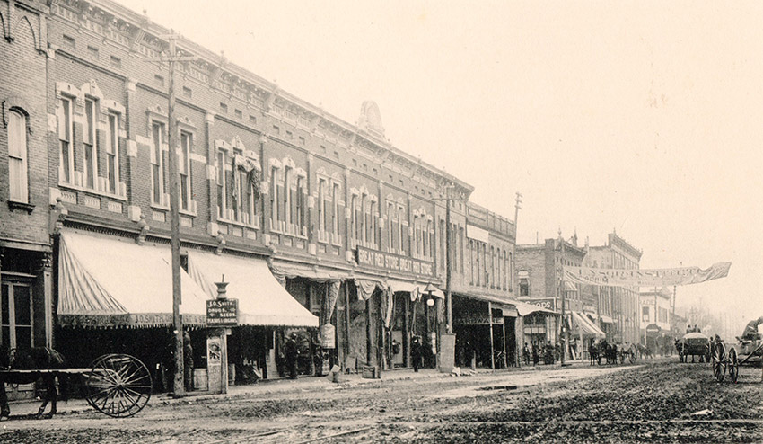 Horse drawn wagons on dirt road with multistory storefronts on one side