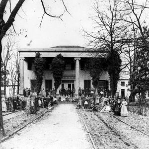 Group of people posing in front of large two-story house with four columns and decorative front garden with rows of trees