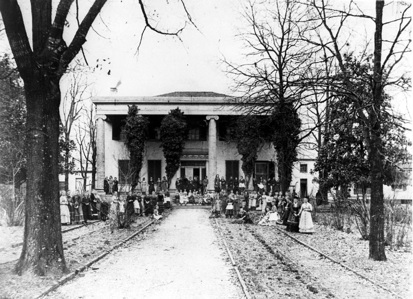 Group of people posing in front of large two-story house with four columns and decorative front garden with rows of trees