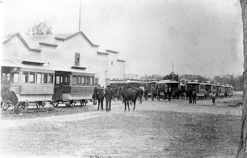 Men with horses and street cars outside a large white building