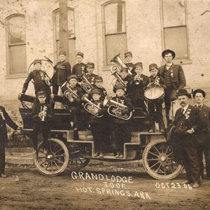 White men in suits and hats standing next to white brass band standing on a wagon outside brick building