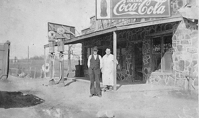 White man and woman standing outside stone storefront with covered porch and gas pumps on dirt road