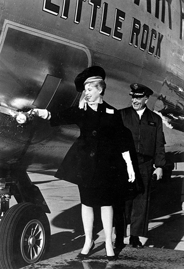 White man in uniform with cap watches white woman in dark dress suit and dark hat breaking bottle on side of large airplane