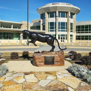Panther statue on rock pedestal with plaque on platform in flower bed and multistory building with round entrance in the background