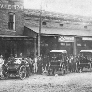 Crowd of white men and women outside brick hardware store and other storefronts with cars on dirt road