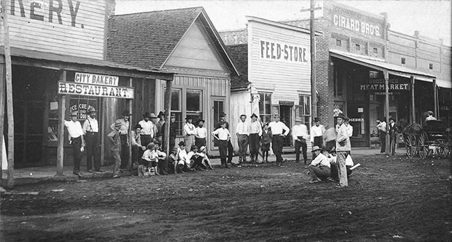 Group of white men in hats on dirt road with storefronts