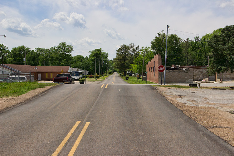 Street with abandoned brick building on right and one-story building with parked cars on left