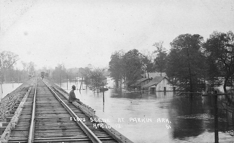 African-American man sitting on railroad bridge during a flood signed "Flood scene at Parkin April 16-17"