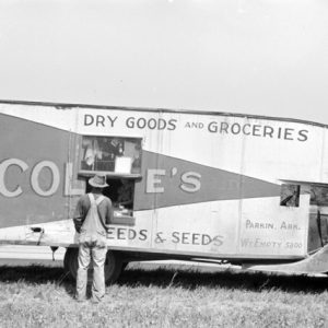 White man standing in a field next to truck displaying the words "dry goods and groceries" with a service window cut in the side