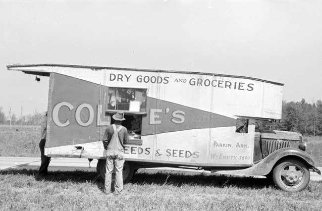 White man standing in a field next to truck displaying the words "dry goods and groceries" with a service window cut in the side