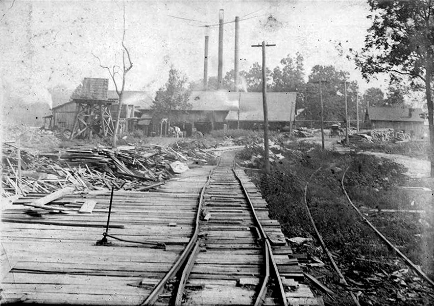 Factory buildings with smokestacks and pile of lumber next to railroad tracks in the foreground