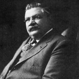 Portrait of a white man seated in a suite and tie with a mustache and wearing glasses.