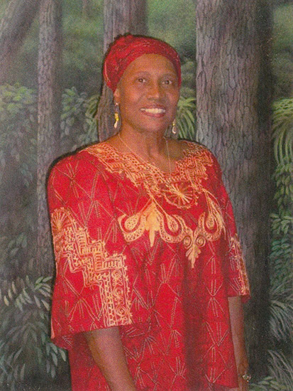 African-American woman smiling in red and gold dress standing with forest background behind her