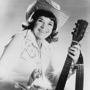 Smiling white woman in western clothing and hat holding an acoustic guitar