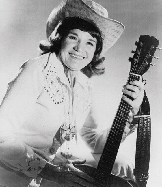 Smiling white woman in western clothing and hat holding an acoustic guitar