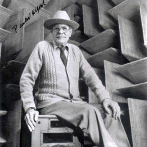 Older white man wearing a hat and glasses and a cardigan sweater sitting on a speaker in a room with sound proofing panels