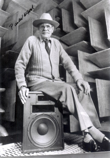 Older white man wearing a hat and glasses and a cardigan sweater sitting on a speaker in a room with sound proofing panels