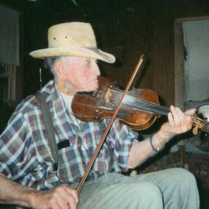 Older white man in hat and shirt with suspenders playing a fiddle indoors