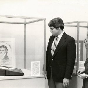 White man and woman looking at picture of white man in glass display case