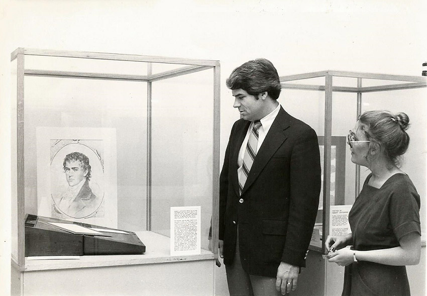 White man and woman looking at picture of white man in glass display case