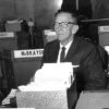 Older white man with glasses in suit sitting at his desk with box full of papers in front of him