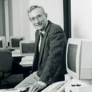 White man with glasses in suit smiling in room with computer on his desk and others on desk behind him