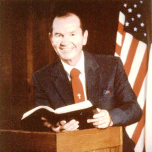 White man in suit and red tie with cross on it at lectern holding book with US flag behind